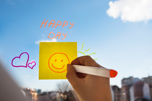Young person drawing smiley face on a sticky note placed on the window. Concept of happiness, motivation, good vibes, positive emotions, success.