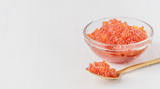 Salty natural salmon red caviar or organic fish roe served in transparent glass bowl on white wooden background with copy space with spoon of gold colour used as spread or ingredient for breakfast