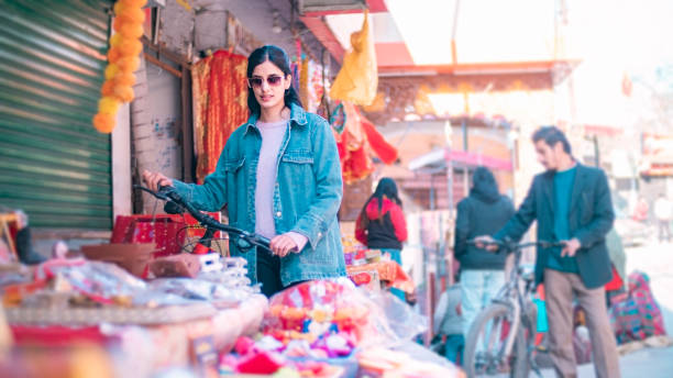 A warm market scene where a smiling young woman draped in a yellow striped dupatta tries on bangles while an older vendor in a pale headscarf assists her. Brightly colored bangles are displayed on rolls and shelves around them, creating a vibrant, textured backdrop that highlights traditional fabrics, jewelry, and a friendly shopping interaction.