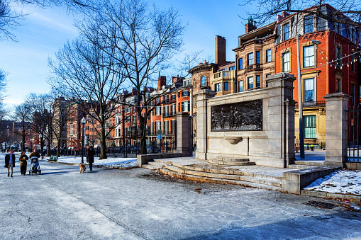 Boston, Massachusetts, USA - January 22, 2024: People walking in Boston Common along Beacon Street and the Beacon Hill neighborhood on a cold winter day. Where Spruce Street meets Beacon Street is the Founders Memorial relief sculpture commemorating the 300th anniversary of the founding of Boston. John Francis Paramino designed it and the city unveiled it in 1930. The sculpture depicts William Blackstone, the city's first European resident, greeting colonial Gov. John Winthrop.