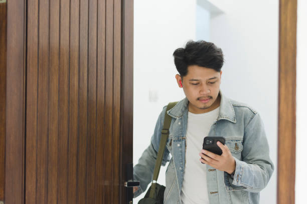 A young Asian man wearing a t-shirt and jeans jacket leaving the house while using a cellphone and earpiece