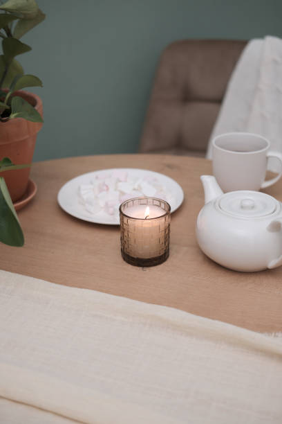 istock Home kitchen interior - white teapot with hot tea, cup of tea, plate with marshmellow, burning candle, green plant in ceramic pot, napkin, beige chair on green background. 1967915092