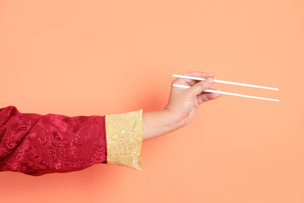 Happy Chinese new year. Hand of asian chinese senior man wearing red traditional cheongsam qipao or changshan dress with gesture of hand holding white chopsticks isolated on orange background.