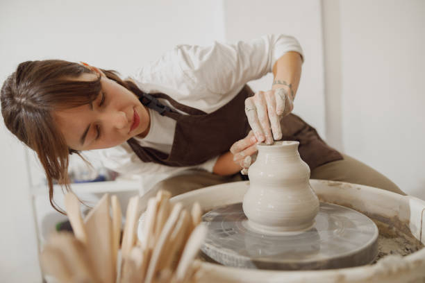 smiling female artisan in apron sitting on bench with pottery wheel and making clay pot - craft zdjęcia i obrazy z banku zdjęć