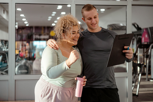 Plus Size, Obese Woman Talking to Fitness Instructor About Her Fitness Program. Overweight, Fat Young Woman in Sportswear Working Out in Gym to Lose Weight. Weight Loss, Healthy Lifestyle