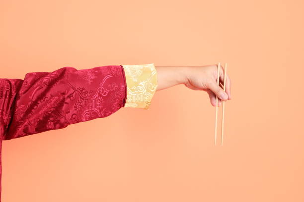 Happy Chinese new year. Hand of asian chinese senior man wearing red traditional cheongsam qipao or changshan dress with gesture of hand holding wood chopsticks isolated on orange background.