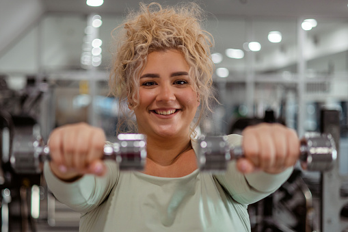 Smiling Chubby Size Woman Looking at Camera While Exercising by Lifting Dumbbell at the Gym. Concept of Weight Loss.