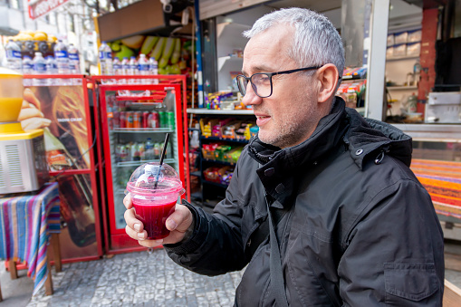 Male tourist drinking a pomegranate juice for refreshment. Portrait face of thirsty  mature solo hipster traveler holding a pomegranate drink on street.