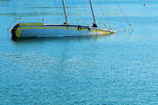 A two mast sailboat lies overturned and half-submerged in tranquil blue waters, with the sails collapsed and draped over the hull.