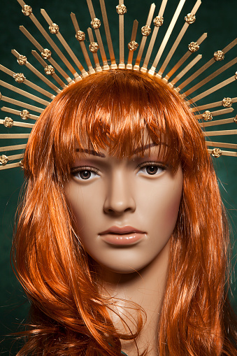 Plastic red haired woman mannequin with bright long hair wearing a spiky golden crown posing on a green background