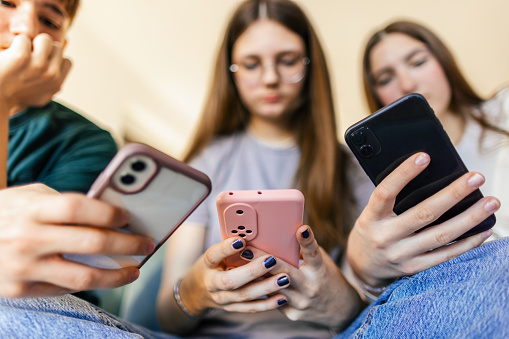 One teenage boy and two teenage girls have fun together using mobile phones at home. Beautiful young people are friends and high school students.
