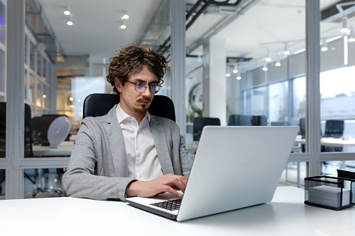 Serious and focused businessman inside modern office working on laptop, bearded man typing on keyboard, thinking man in business clothes sitting at workplace.
