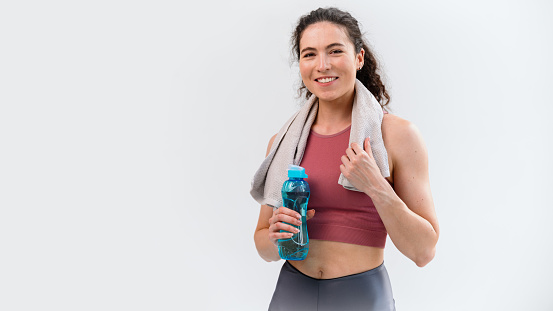 Banner view of sporty smiling woman in sportswear and white towel on neck holding blue plastic bottle with healthy drink, fresh water and looking at camera. Copy space