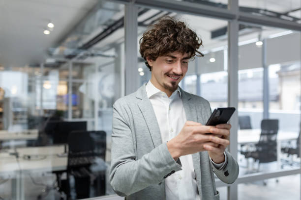 istock Successful businessman with beard standing near window, man using phone, happy boss typing message and browsing online pages on smartphone app inside office. 1968101336