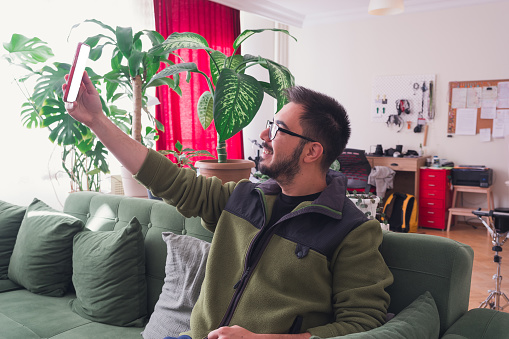 Seated comfortably on the couch at home, a man smiles as he takes a selfie with his smartphone. The white mockup screen reflects the joy captured in the moment, blending modern technology with the warmth of home.
