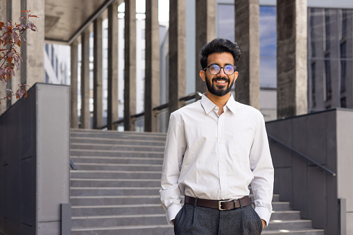 Confident young businessman smiling outdoors with modern architecture, wearing a white shirt and glasses.