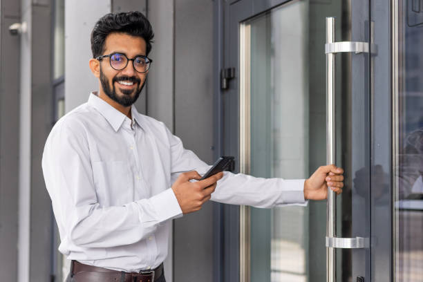 istock Smiling professional man with glasses using smartphone while entering modern office building, business casual 1968124140