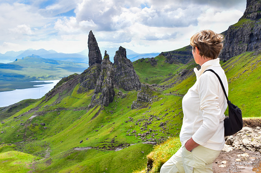 Woman dressed in white looking at the Old Man of Storr rock formation landscape and enjoying the amazing view. Isle of Skye in summer, Scotland landmark, UK, travel Europe.