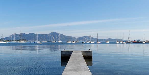 A tranquil concrete pier stretches out into a calm blue bay, where a flotilla of sailboats peacefully anchors against a backdrop of gentle hills under a clear sky. The mirror-like water reflects the boats and distant mountains, creating a scene of perfect symmetry and stillness