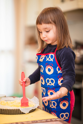 A young girl in an apron prepares a cake with a serious expression, brushing some ingredients on the top of the cake with an appropriate brush.