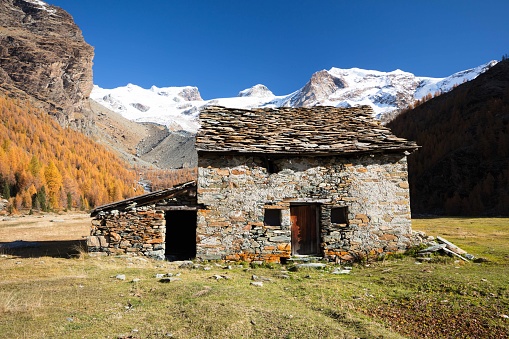 The majestic peaks of Monte Rosa, on a sunny, crystal-clear autumn morning, stand out against the backdrop of a vast glacial valley that has become an alpine pasture for centuries. A crumbling shepherds' hut stands in the center of the plain, poignantly empty and lonely.