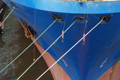 Head lines coming out from bow of container vessel with blue hull and anchor and passing through panama bow bulwark and other other bow deck mounted with rollers. On the ropes are wooden rat guards.