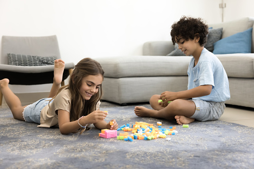 Two cute happy sibling kids stacking toy building blocks on warm carpeted floor, playing learning creative game at home, enjoying friendship, childhood, smart activity, playtime