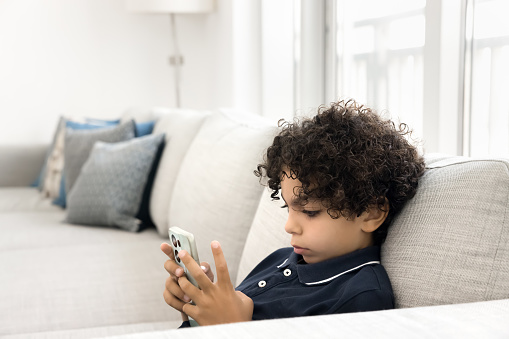 Cute Latin boy playing learning game on mobile phone, using online educational application on smartphone, browsing Internet with safe access, resting on spacious sofa at home,