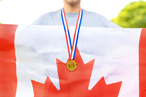 Triumphant Canadian male athlete proudly showcases a gold medal, symbolizing victory. Holding the Canadian flag, he embodies national pride during sporting events.