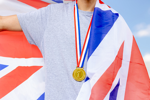 Triumphant British male athlete proudly showcases a gold medal, symbolizing victory. Holding the Union Jack flag, he embodies national pride during sporting events.