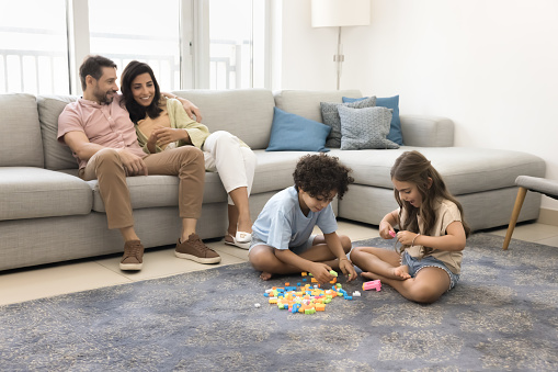 Two cute engaged kids playing with toy blocks on carpeted floor, constructing colorful cubes, enjoying learning game with heap of plastic pieces. Mom and dad watching little children
