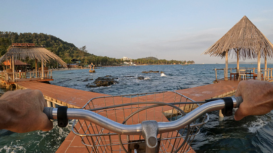 Personal perspective of man riding bicycle along pier over a calm bay with fork in pathway