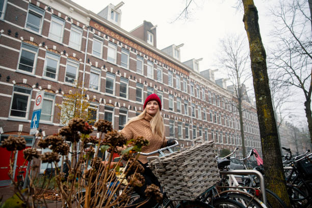 Amsterdam, Holland: A woman carries twin dogs in her bike “basket” at a busy sunny intersection in downtown Amsterdam, as another woman passes on a bike in the foreground, and a crowd gathers at a coffee bar in the background.