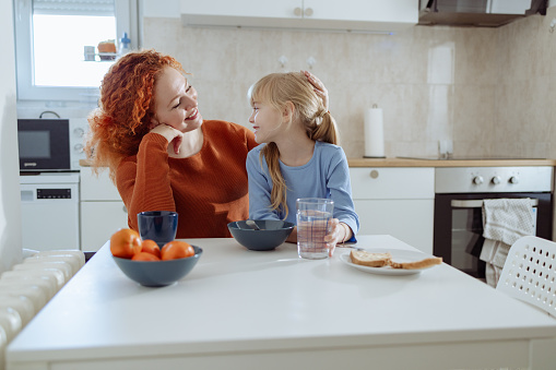 Beautiful mother is with her little daughter in the kitchen. They are talking and enjoying breakfast