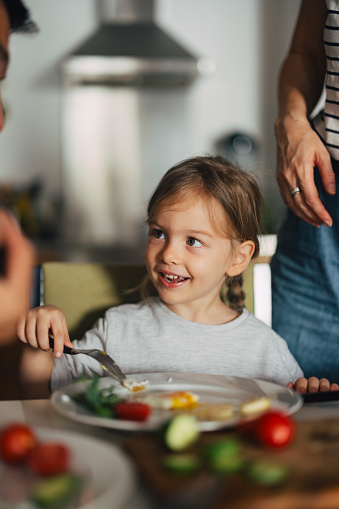 Close up shot of a sweet little girl sitting at the table and eating her breakfast in the morning. She is smiling and looking away.