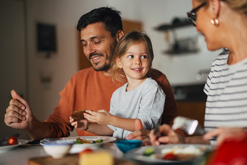 Portrait of a cute young daughter sitting together with her happy parents and eating breakfast in the morning at home.