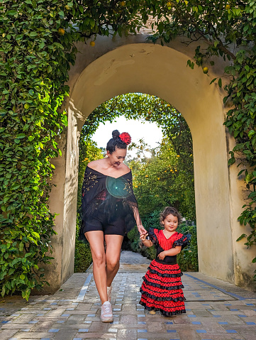 Beautiful mother and young daughter dressed in red flamenco dress walking holding hands atthe Royal Alcazar gardens, Seville, Spain