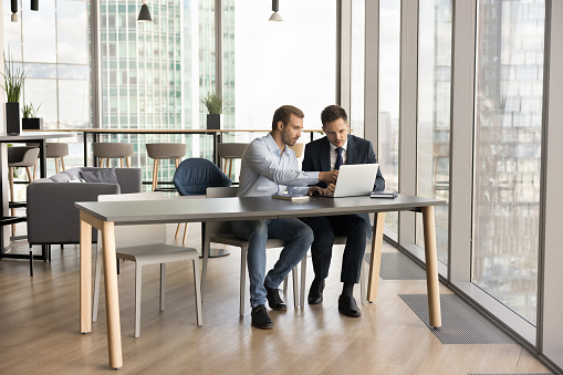 Two business colleagues men working at laptop in modern office interior, talking at workplace table in co-working space, discussing online startup project in urban workspace with large window