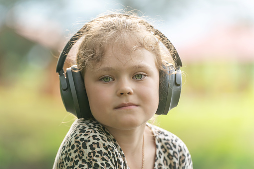 Cute little girl in modern wireless headphones enjoys music outdoors.