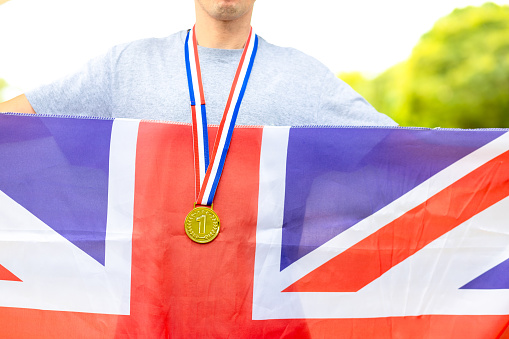 Triumphant British male athlete proudly showcases a gold medal, symbolizing victory. Holding the Union Jack flag, he embodies national pride during sporting events.