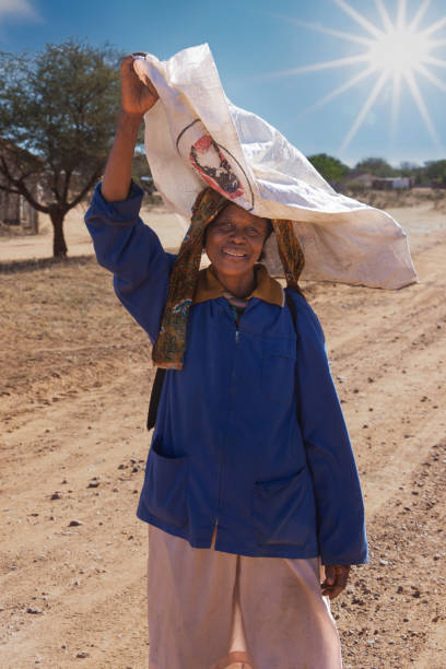 mulher africana idosa, carregar um saco em sua cabeça, andando em uma estrada de terra na aldeia - african culture farmer action activity - fotografias e filmes do acervo