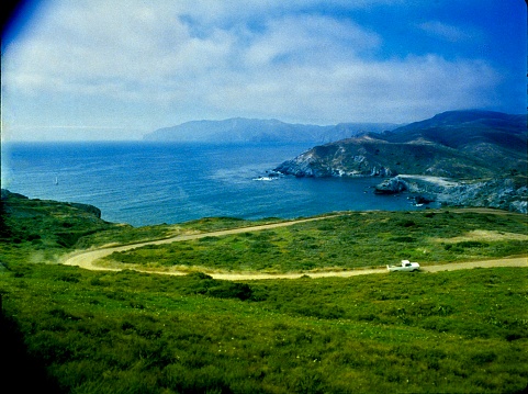 This photograph captures a serene coastal landscape with a winding dirt road leading to a secluded bay, surrounded by lush greenery and rolling hills under a hazy blue sky.
