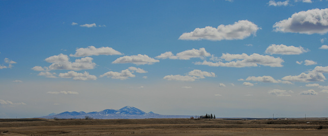 A photo of the Sweetgrass Hills in Montana, USA taken from the south edge of Milk River, Alberta.  There are white, puffy clouds in a blue sky, and a low haze.  Although this appears to be one mass, it is actually a combination of separate hills.  As a group, their appearance changes as the location one is viewing from moves.