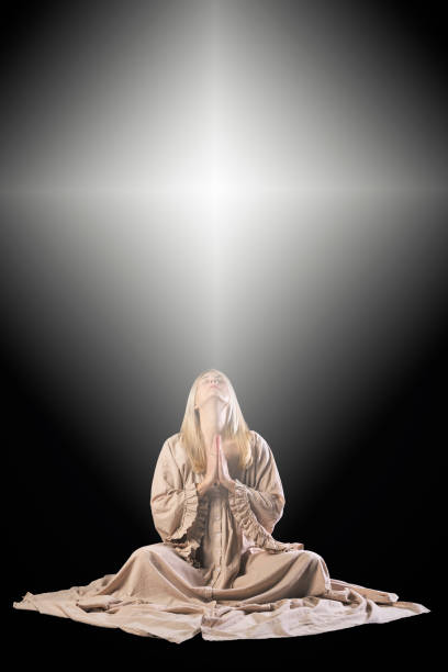 A woman sitting in prayer on a black background with an illuminated cross overhead.
