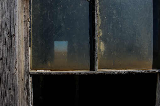 Photo looks through both the glassed window in the front of the house, and the empty window in the back of the house.  A view of sky, prairie and a river is seen through the back window.  The front window has intact dirty glass in the upper section of the window.  An old wood house in Southern Alberta, Canada.  Built in the early 1900’s and abandoned for decades, there is extensive wear showing.  This house was built for settlers who moved up from the United States.  It was torn down in 2008 to make room for an expanded highway.