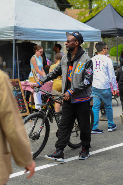 Stylish African American man wearing sunglasses and bomber jacket, pushing his bicycle through a summer afternoon farmer's market in May 2023, in the city of Kingston New York, shopping for fresh local farm foods and crafts from local artisans.
