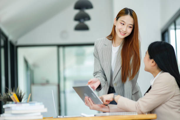 istock Office woman is standing and teaching a new employee. It describes the work format displayed on the tablet screen. 1968776954