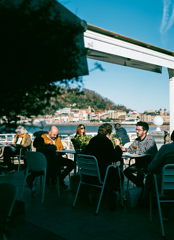 1st January 2024, San Sebastián，Basque Country, Spain: Locals and Tourists enjoying cafe by beach on a sunny day. San Sebastián is famous for its longest beach in Europe and incredible food culture. The cityscape can be seen in the background.