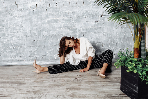 Caucasian woman in her 30s, elegantly seated on a wooden floor, wearing a white blouse and striped pants. Front view angle with a green plant. Concept: fashion and interior design inspiration.