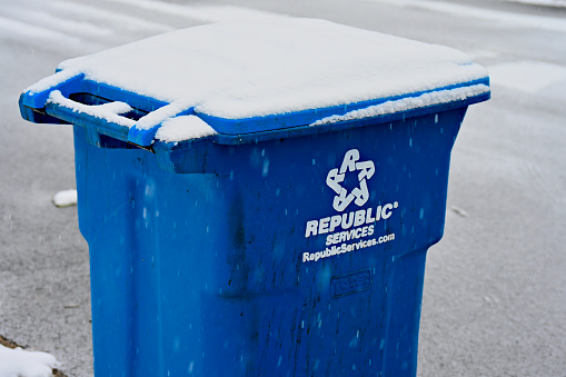 Fairfax, Virginia, USA - January 15, 2024: Falling snow covers a Republic Services recycling bin and the street during a heavy snow storm in Northern Virginia.